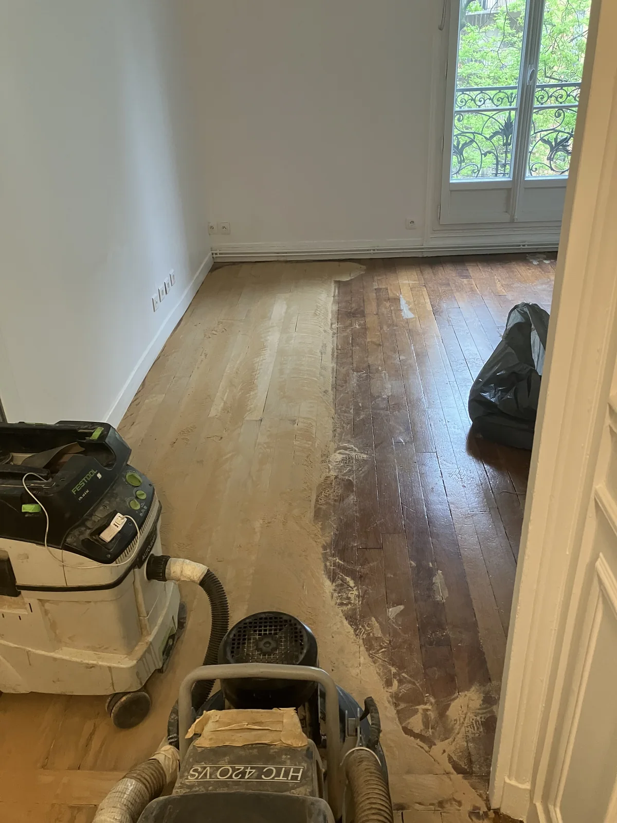 À droite&nbsp;: le parquet tel que je l'ai trouvé — chêne noirci par des décennies de vernis, taches, lames marquées. À gauche&nbsp;: le même bois, après le passage de la machine. La machine planétaire HTC 420 VS et l'aspirateur Festool CTL 48 E sont au premier plan. Aucune retouche, aucun décor&nbsp;: c'est la limite réelle entre l'avant et l'après.