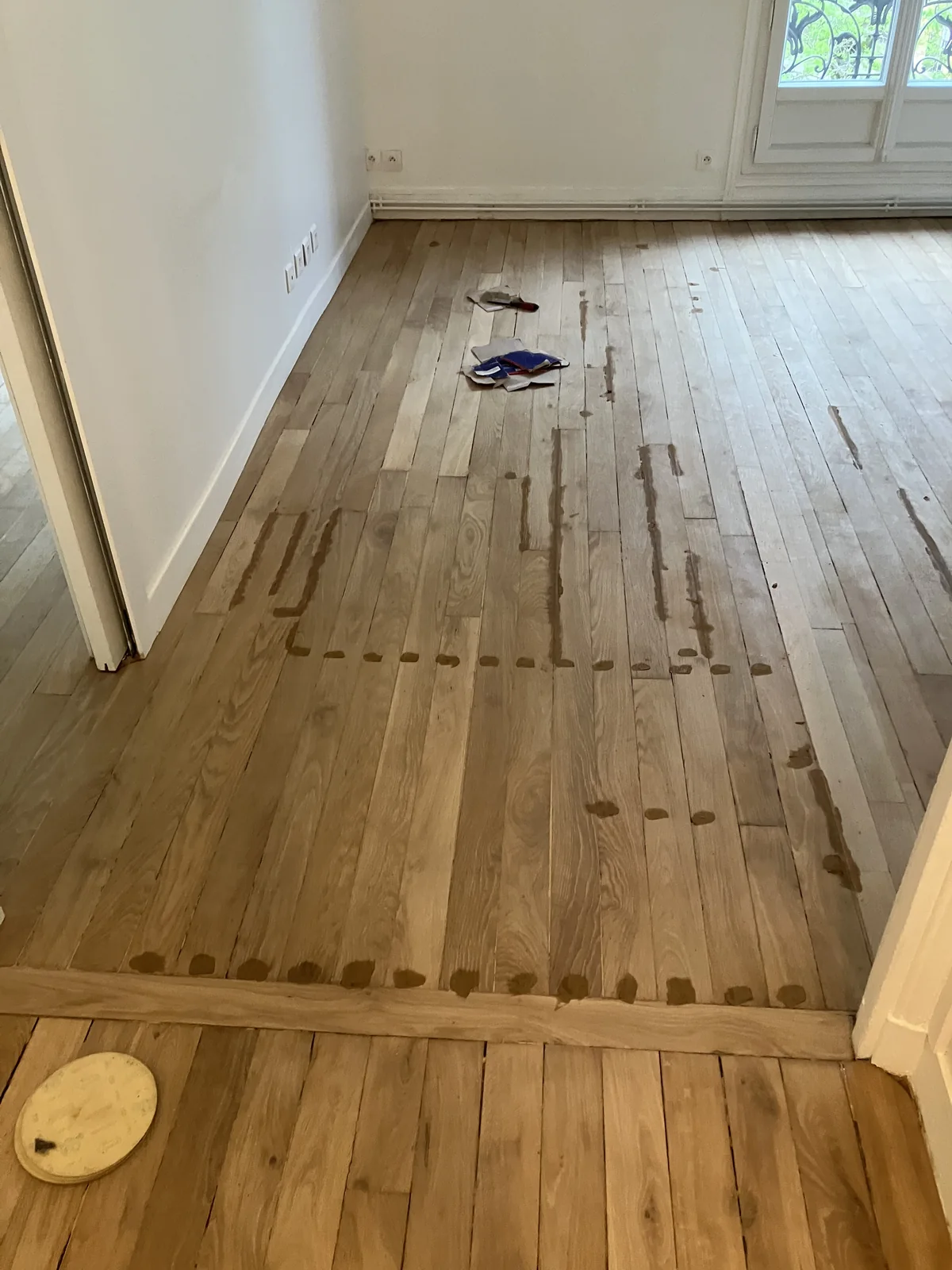 Le parquet est poncé, mais pas encore vitrifié. On voit les points sombres de pâte à bois Sintobois&nbsp;: trous de clous, fissures, joints trop larges rebouchés un par un. Cette résine bi-composant est teintable et souple — elle ne fissure pas quand le bois travaille. Le rebouchage se fait <strong>toujours avant</strong> la vitrification, jamais après. Une fois le vernis passé, ces réparations deviennent invisibles.